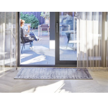 Rechteckiger Teppich mit grauem Holzplankenmuster und dekorativer Bordüre in einem hellen Wohnzimmer vor einer Terrassenschiebetür.