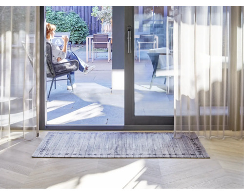 Rechteckiger Teppich mit grauem Holzplankenmuster und dekorativer Bordüre in einem hellen Wohnzimmer vor einer Terrassenschiebetür.