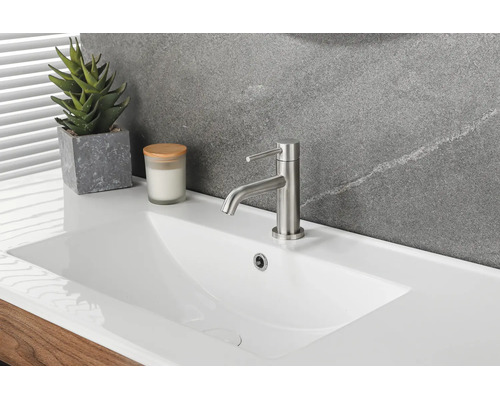Modernes weißes Keramik-Waschbecken mit einer Einhand-Waschtischarmatur aus gebürstetem Metall, einer Sukkulente und Kerze vor einer Steinwand.