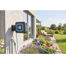 Wand-Schlauchbox mit for.q Logo an einer Hauswand montiert in einem Garten mit Blumenbeeten, Wasseranschluss, Terrasse und Rasenfläche.