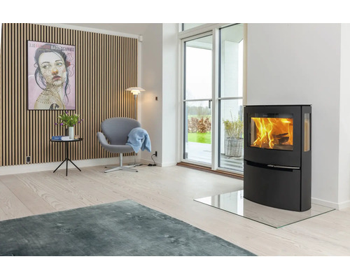 Modernes Wohnzimmer mit schwarzem Kaminofen auf Glasplatte, Holzlamellenwand, grauem Sessel und großer Fensterfront zum Garten.