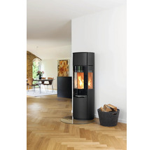 Moderner schwarzer Kaminofen mit brennendem Feuer auf einer Glasbodenplatte neben einem Holzkorb in einem hellen Wohnzimmer mit Parkettboden.