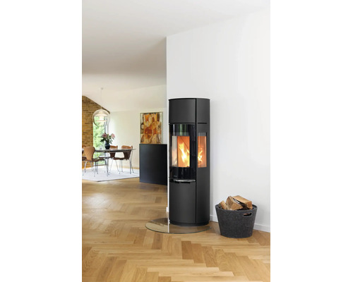 Moderner schwarzer Kaminofen mit brennendem Feuer auf einer Glasbodenplatte neben einem Holzkorb in einem hellen Wohnzimmer mit Parkettboden.