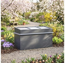Anthrazitfarbenes Frühbeet aus Metall mit durchsichtigem Deckel in einem frühlingshaften Garten mit Blumen und blühenden Bäumen.