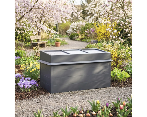 Anthrazitfarbenes Frühbeet aus Metall mit durchsichtigem Deckel in einem frühlingshaften Garten mit Blumen und blühenden Bäumen.