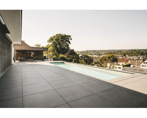 Moderne Dachterrasse mit großformatigen grauen Steinfliesen und einem schmalen Infinity-Pool. Im Hintergrund ist eine Lounge. Helle Atmosphäre.