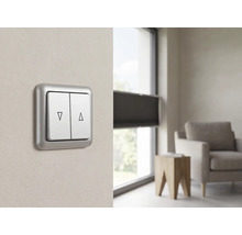 Doppelwipp-Rollladenschalter mit Auf- und Ab-Symbolen an einer Wand in einem modernen, hellen Wohnzimmer neben einem Fenster mit Jalousien.
