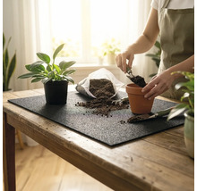 Person beim Umtopfen einer Zimmerpflanze auf einer schwarzen Gartenmatte mit Erde, Tontopf und Handschaufel auf einem Holztisch.