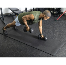 Ein Mann führt Krafttraining mit Hanteln auf einer schwarzen Fitnessmatte in einem hellen, organisierten Fitnessraum mit Gummiboden aus.