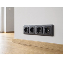 Schwarze 4-fach Schutzkontakt-Steckdose an einer hellen Wand über einer Fußleiste aus Holz in einem hellen, modern gestalteten Raum.