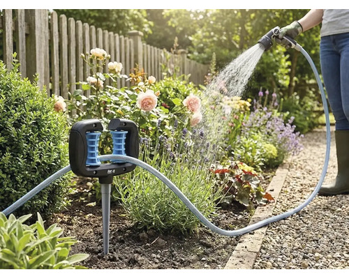 Schlauchführung in einem Blumenbeet mit einem Gartenschlauch und einer Person, die im Hintergrund Pflanzen gießt.