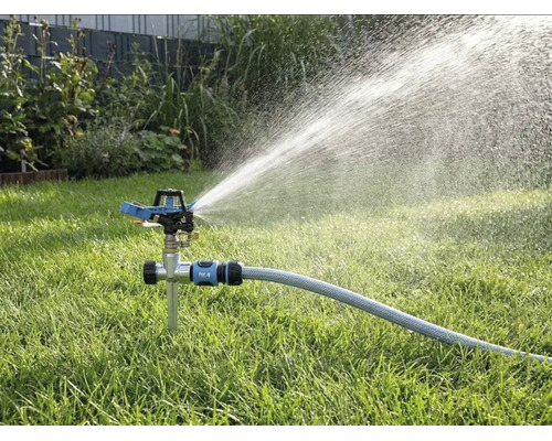 Impulsregner mit Erdspieß auf einer Rasenfläche, angeschlossen an einen Gartenschlauch, sprüht Wasser in einem weiten Strahl.