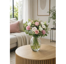 Blumenstrauß mit Rosen und Nelken in einer Glasvase auf einem runden Holztisch in einem hellen Wohnzimmer.