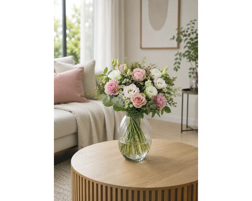 Blumenstrauß mit Rosen und Nelken in einer Glasvase auf einem runden Holztisch in einem hellen Wohnzimmer.