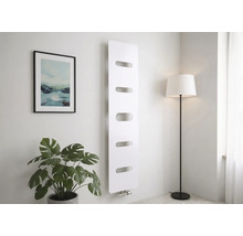Moderner Vertikalheizkörper in Weiß mit fünf horizontalen Aussparungen, montiert an einer hellen Wand neben einer Zimmerpflanze und Stehlampe.