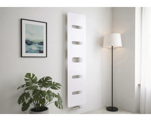 Moderner Vertikalheizkörper in Weiß mit fünf horizontalen Aussparungen, montiert an einer hellen Wand neben einer Zimmerpflanze und Stehlampe.