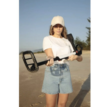 Eine Frau hält einen Metalldetektor mit einer rechteckigen Suchspule an einem Sandstrand in den Händen.