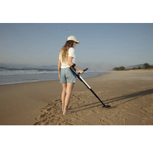 Eine Frau verwendet einen Metalldetektor an einem weitläufigen Sandstrand am Meer.