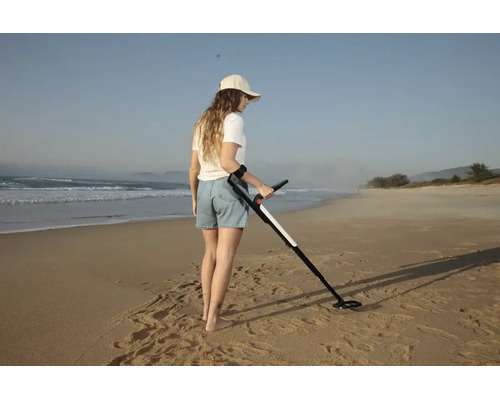 Eine Frau verwendet einen Metalldetektor an einem weitläufigen Sandstrand am Meer.