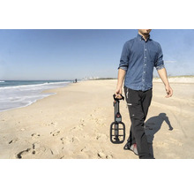 Ein Mann geht am Strand spazieren und hält einen kompakten Metalldetektor in der Hand. Im Hintergrund sind das Meer und Wellen zu sehen.