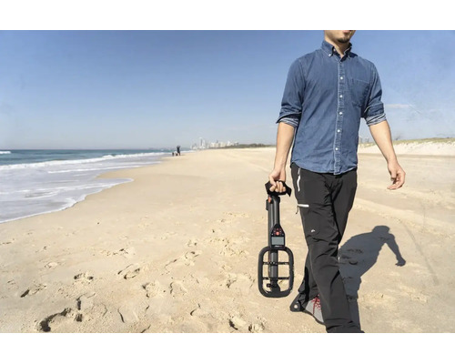Ein Mann geht am Strand spazieren und hält einen kompakten Metalldetektor in der Hand. Im Hintergrund sind das Meer und Wellen zu sehen.