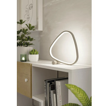 Moderne, dreieckige LED-Tischleuchte auf einem weißen Regal neben grünen Pflanzen und Büchern in einem hellen, minimalistischen Raum.