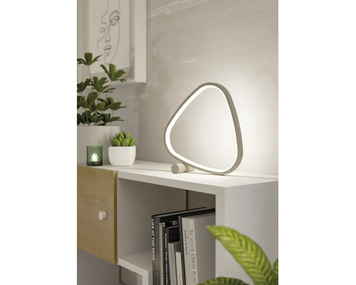 Moderne, dreieckige LED-Tischleuchte auf einem weißen Regal neben grünen Pflanzen und Büchern in einem hellen, minimalistischen Raum.