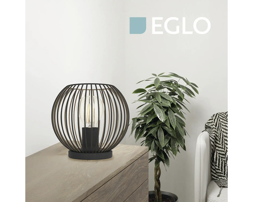 Moderne Eglo Tischleuchte aus Metall im Gitterdesign auf einer Holzkommode neben einer Zimmerpflanze in einem hellen Raum. Eglo Logo.