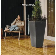 Hoher, konischer Pflanzkübel in Anthrazit auf einer Holzterrasse neben einer sitzenden Person. Tierra Verde Logo.