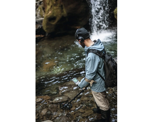 Eine Person mit Rucksack und Kopfhörern verwendet einen Metalldetektor in einem flachen, steinigen Bachlauf vor einem Wasserfall.
