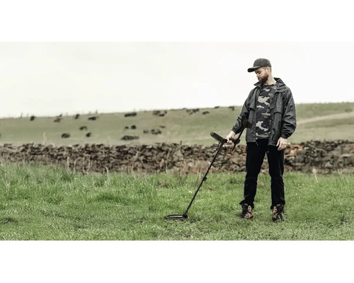 Ein Mann sucht mit einem Metalldetektor auf einer Wiese nach Gegenständen vor einer Steinmauer.