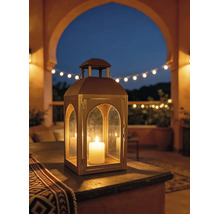 Metalllaterne mit Rundbogen-Glasfenstern und brennender Kerze auf einer Terrasse bei Dämmerung, mit Lichterkette im Hintergrund.