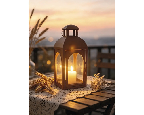 Laterne mit brennender Kerze auf einem Holztisch mit Spitzendeckchen, Weizen und Trockenblumen vor einem stimmungsvollen Sonnenuntergang.