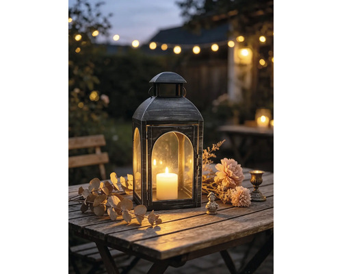 Metalllaterne mit brennender Stumpenkerze auf einem Holztisch im Garten, dekoriert mit Trockenblumen und Blättern.