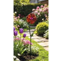 Dekorativer Gartenstecker aus Metall in Form einer orangefarbenen Blume in einem hellen Gartenbeet mit Tulpen und einem Steinpfad.