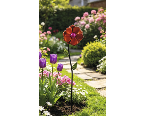 Dekorativer Gartenstecker aus Metall in Form einer orangefarbenen Blume in einem hellen Gartenbeet mit Tulpen und einem Steinpfad.