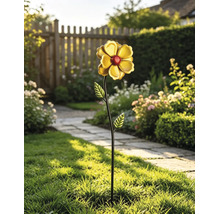 Dekorativer Metall-Gartenstecker als Blume mit gelben Blättern und roter Mitte auf Rasen in hellem Garten mit Steinweg und Holzzaun.