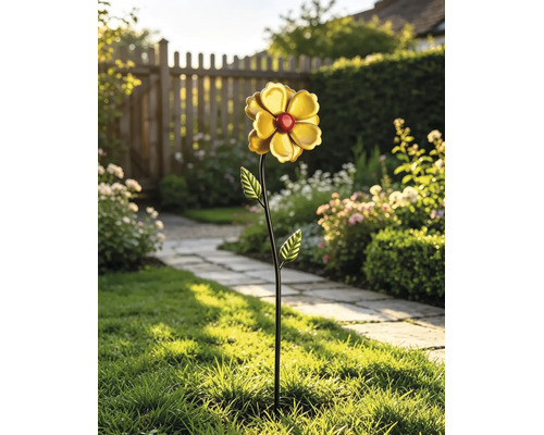 Dekorativer Metall-Gartenstecker als Blume mit gelben Blättern und roter Mitte auf Rasen in hellem Garten mit Steinweg und Holzzaun.