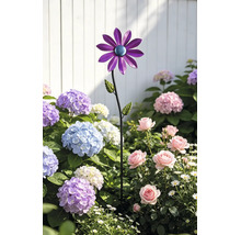 Gartenstecker aus Metall in Blumenform mit lila Blütenblättern in einem Beet mit Hortensien und Rosen vor einem weißen Zaun.