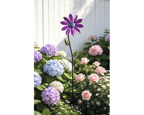 Gartenstecker aus Metall in Blumenform mit lila Blütenblättern in einem Beet mit Hortensien und Rosen vor einem weißen Zaun.