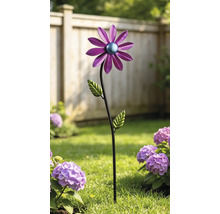 Dekorativer Gartenstecker aus Metall in Blumenform mit lila Blüte und grünen Blättern in einem Gartenbeet vor einem Holzzaun.