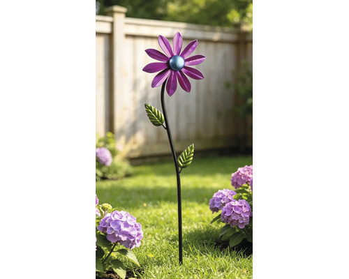 Dekorativer Gartenstecker aus Metall in Blumenform mit lila Blüte und grünen Blättern in einem Gartenbeet vor einem Holzzaun.