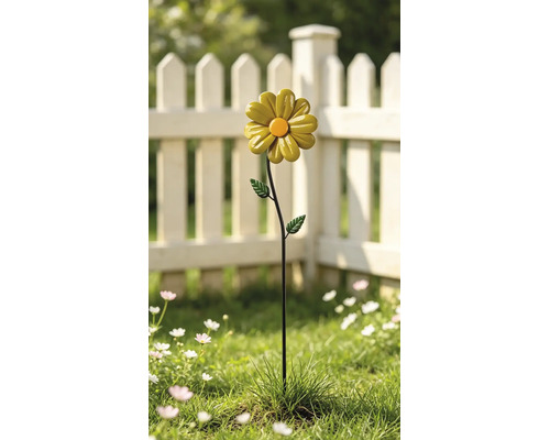 Gartenstecker aus Metall in Form einer gelben Blume mit orangefarbener Mitte in einem Garten mit weißem Lattenzaun.