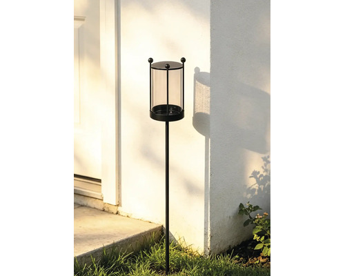 Gartenfackel mit Glaseinsatz und schwarzem Metallgestell auf einem Erdspieß vor einer weißen Hauswand.