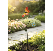 Gartenstecker aus Metall mit Schmetterling und Blume in einem Gartenbeet neben einem gepflasterten Weg.