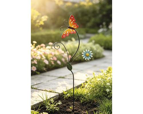 Gartenstecker aus Metall mit Schmetterling und Blume in einem Gartenbeet neben einem gepflasterten Weg.