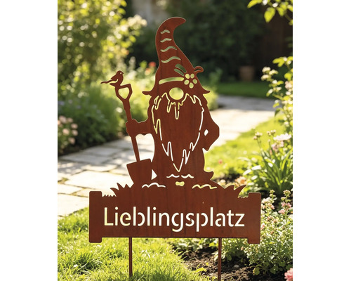 Rostiger Gartenstecker in Form eines Wichtels mit Schaufel und dem Schriftzug Lieblingsplatz, stehend in einem bepflanzten Garten.
