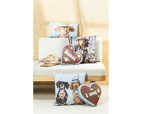 Verschiedene Dekokissen mit bayerischen Motiven wie Dackeln, Kühen und Lebkuchenherzen auf einem Sofa vor einer Holzwand.