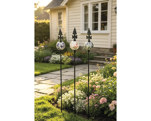 Drei dekorative Gartenstäbe aus Metall mit verzierten Keramikkugeln und Lilien-Spitzen in einem blühenden Gartenbeet.