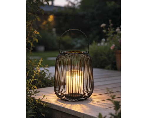 Schwarze Metalldrahtlaterne mit Glaseinsatz und brennender Kerze auf einer Holzterrasse im Garten.